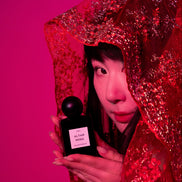 Person holding a black perfume bottle with a red veil and pink background