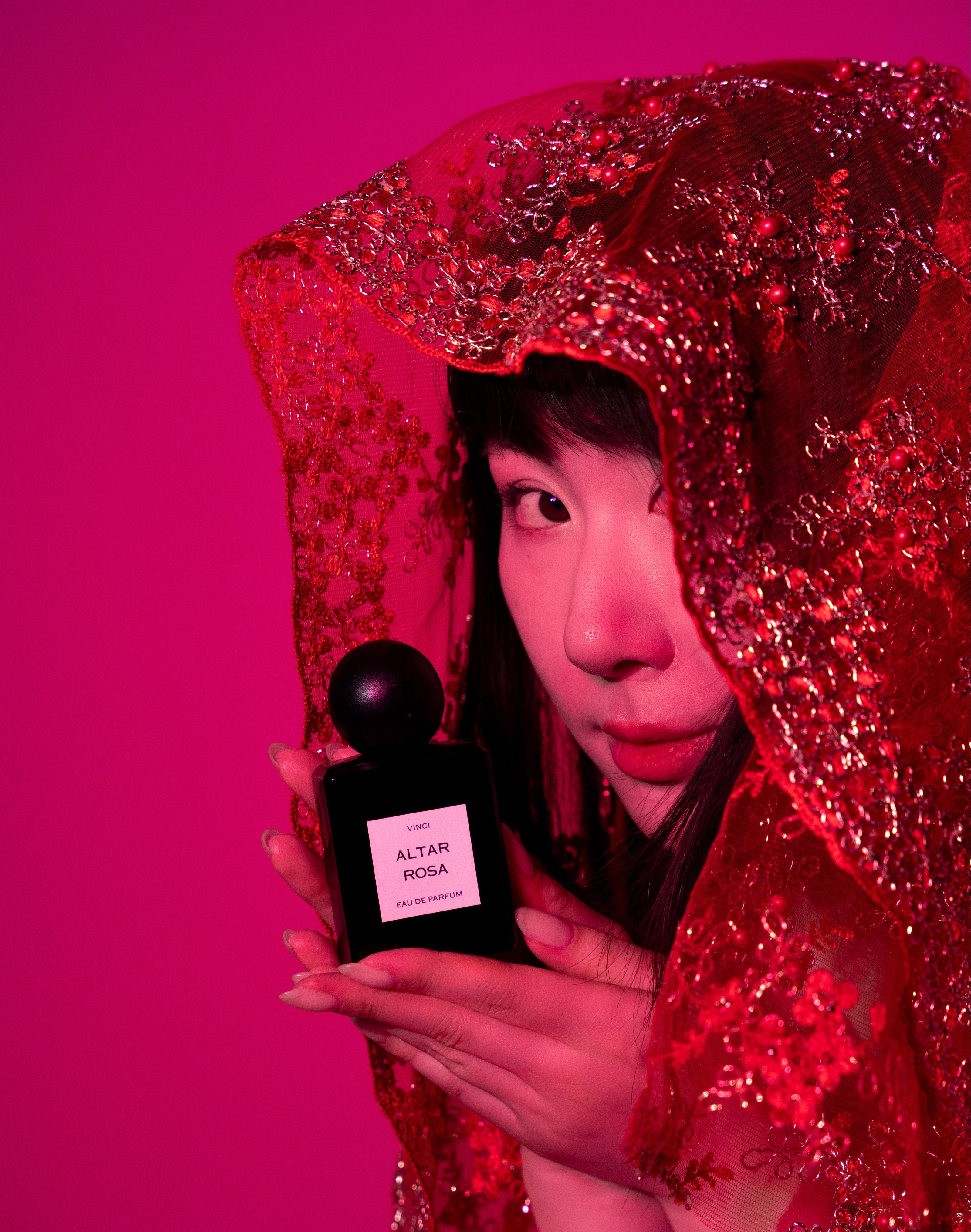 Person holding a black perfume bottle with a red veil and pink background